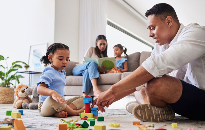 Father playing with young daughter on the floor while mom and other daughter are reading a book on the couch.